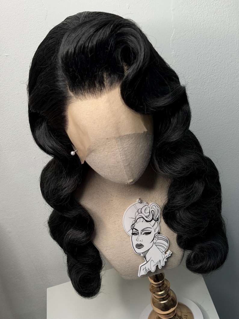 May include: A black wig with voluminous, wavy curls styled in a vintage Hollywood fashion. The wig is displayed on a mannequin head with a lace front. A small tag with a graphic of a woman's face is attached to the mannequin.
