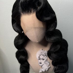 May include: A black wig styled with vintage waves, displayed on a mannequin head. The wig features a lace front and is adorned with a small pearl earring. A sticker with a graphic of a woman's face is attached to the mannequin.