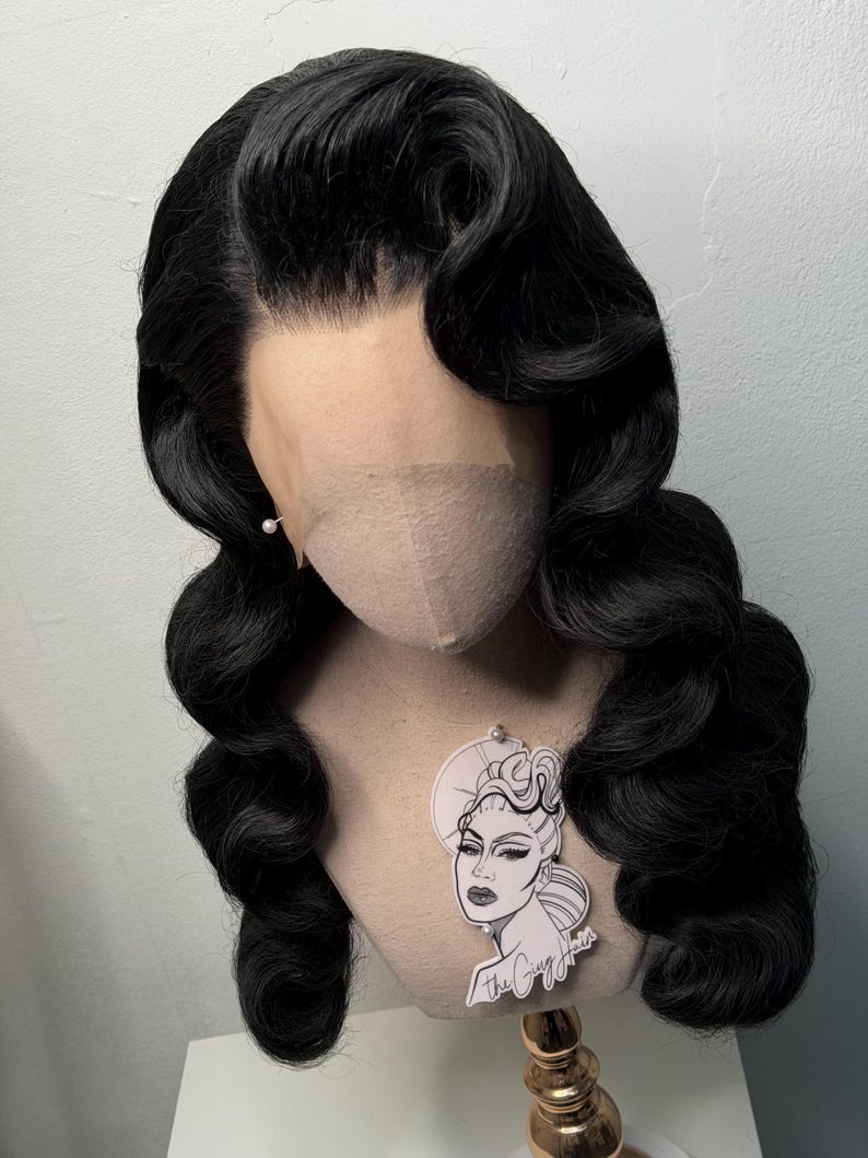 May include: A black wig styled with vintage waves, displayed on a mannequin head. The wig features a lace front and is adorned with a small pearl earring. A sticker with a graphic of a woman's face is attached to the mannequin.