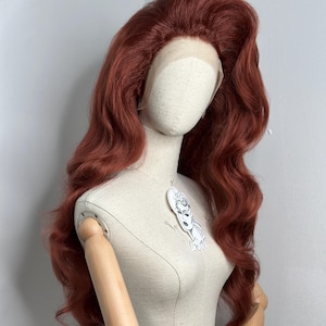 Peut inclure: Perruque auburn avec de longues vagues volumineuses, présentée sur un mannequin. La perruque a un devant en dentelle et une coiffure haute. Le mannequin a des bras en bois et un torse crème. Une petite illustration est épinglée sur le mannequin.