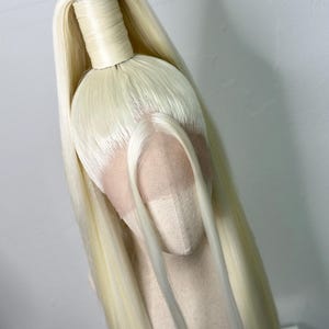 Könnte beinhalten: Eine lange, platinblonde Perücke, die zu einem hohen Pferdeschwanz mit einem Stoffhaargummi gebunden ist. Die Perücke befindet sich auf einem Mannequin-Kopf.