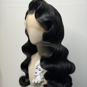 May include: A black, wavy wig styled in a vintage, glamorous fashion. The wig is placed on a mannequin head with a light skin tone. The hair is styled with large, defined waves and a side part. A sticker with a woman's face is attached to the mannequin.
