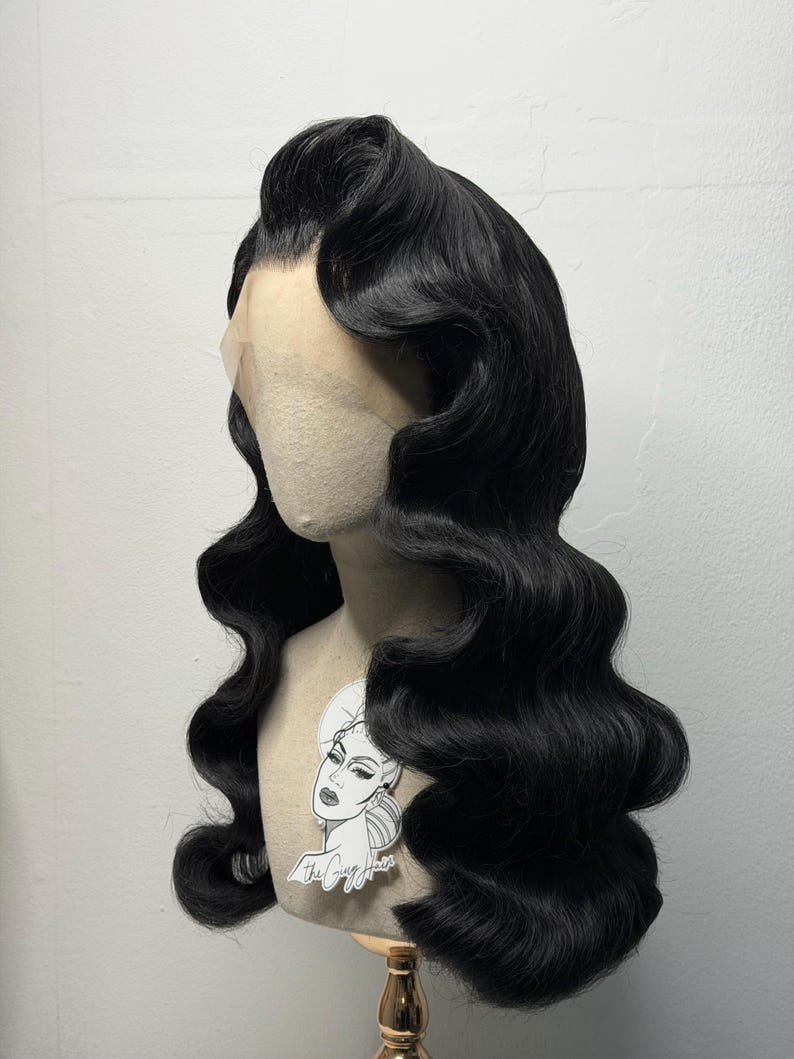 May include: A black, wavy wig styled in a vintage, glamorous fashion. The wig is placed on a mannequin head with a light skin tone. The hair is styled with large, defined waves and a side part. A sticker with a woman's face is attached to the mannequin.