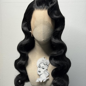 May include: A black, wavy wig styled in a vintage Hollywood fashion, displayed on a mannequin head. The wig features a deep side part and voluminous curls. A small, decorative tag with a woman's portrait hangs from the mannequin.