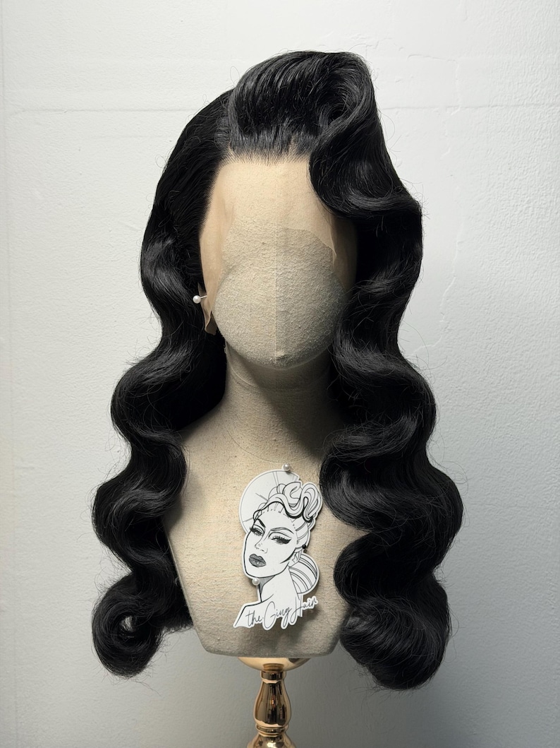 May include: A black, wavy wig styled in a vintage Hollywood fashion, displayed on a mannequin head. The wig features a deep side part and voluminous curls. A small, decorative tag with a woman's portrait hangs from the mannequin.