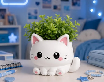 Puede incluir: Una maceta blanca con forma de gato con detalles rosas, que contiene una planta verde vibrante. La maceta presenta un diseño lindo con ojos negros, bigotes y mejillas rosas. Una adición encantadora a cualquier habitación.