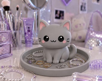 Puede incluir: Una figura de gato gris con grandes ojos negros y detalles en rosa se encuentra sobre una bandeja circular gris. La bandeja contiene varias piezas de joyería, incluyendo anillos, pulseras y collares, dispuestos alrededor de la figura.
