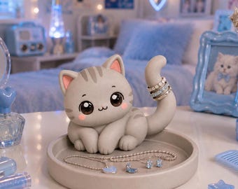 Puede incluir: Un soporte para joyas con forma de gato gris, grandes ojos negros y mejillas rosadas, sobre una bandeja circular. La cola del gato está curvada hacia arriba para sostener anillos, mientras que la bandeja muestra un collar de cadena plateada.