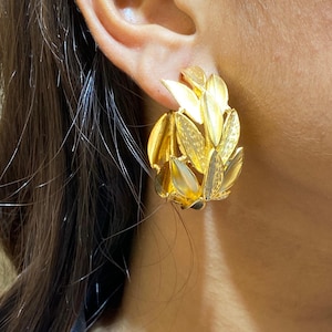 May include: A gold-toned hoop earring featuring a cluster of layered, textured leaf-shaped elements. The leaves exhibit a mix of polished and brushed finishes, creating a dimensional, botanical-inspired design that curves around the earlobe.