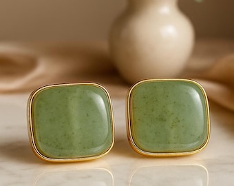 Könnte beinhalten: Ein Paar quadratische Ohrstecker mit glatten, hellgrünen Steincabochons mit feinen dunklen Sprenkeln, jeweils eingefasst in einen polierten goldfarbenen Metallrahmen. Die Ohrringe haben eine glänzende Oberfläche und abgerundete Ecken.