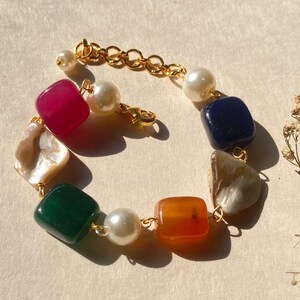 Könnte beinhalten: Ein farbenfrohes Armband mit einer Vielzahl von Edelsteinen und Perlen. Das Armband umfasst quadratische und unregelmäßig geformte Steine in Rosa, Grün, Orange, Blau und neutralen Tönen, verbunden durch goldfarbene Kettenglieder.