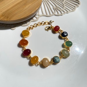Könnte beinhalten: Ein goldenes Kettenarmband mit ovalen Edelsteinen in verschiedenen Farben, darunter Rot, Orange, Gelb, Grün, Blau und Schwarz. Das Armband hat einen Karabinerverschluss.