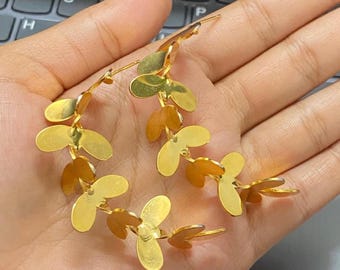 May include: A pair of gold-toned earrings featuring a curved, branch-like design composed of multiple smooth, reflective, oval-shaped leaf elements. The earrings have a polished metallic finish and a post-back closure mechanism.