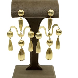 Puede incluir: Un par de pendientes tipo candelabro en tono dorado con una serie de componentes acanalados, esféricos y en forma de lágrima suspendidos de un marco de metal curvo. El diseño incluye múltiples elementos colgantes con textura.