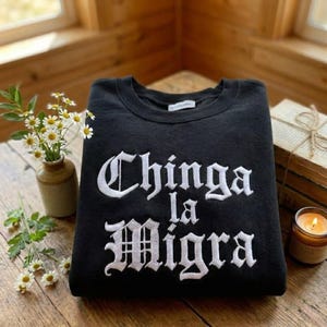 Könnte beinhalten: Schwarzes Sweatshirt mit weiß gesticktem Text "Chinga la Migra". Das Rundhals-Sweatshirt ist auf einer Holzoberfläche gefaltet. Eine kleine Vase mit Gänseblümchen, ein Stapel Bücher und eine brennende Kerze befinden sich im Hintergrund.