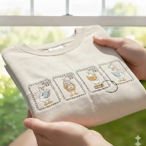 May include: Beige crewneck sweatshirt featuring four postage stamp designs with cartoon ducks. Each stamp has a different duck illustration, holding books or mail. The stamps are outlined with a decorative border and the number "33".