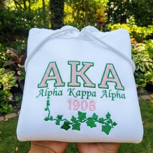 Puede incluir: Sudadera con capucha blanca con las letras "AKA" en verde con contornos rosas. Debajo, las palabras "Alpha Kappa Alpha" y el número "1908" en rosa. Diseño de hiedra verde en la parte inferior.