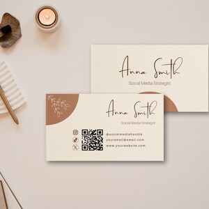 May include: A beige business card with brown accents. The card features the name "Anna Smith" in cursive script, followed by the title "Social Media Strategist". The card also includes a QR code, social media icons, and contact information: @socialmediahandle, youremail@email.com, and www.yourwebsite.com.