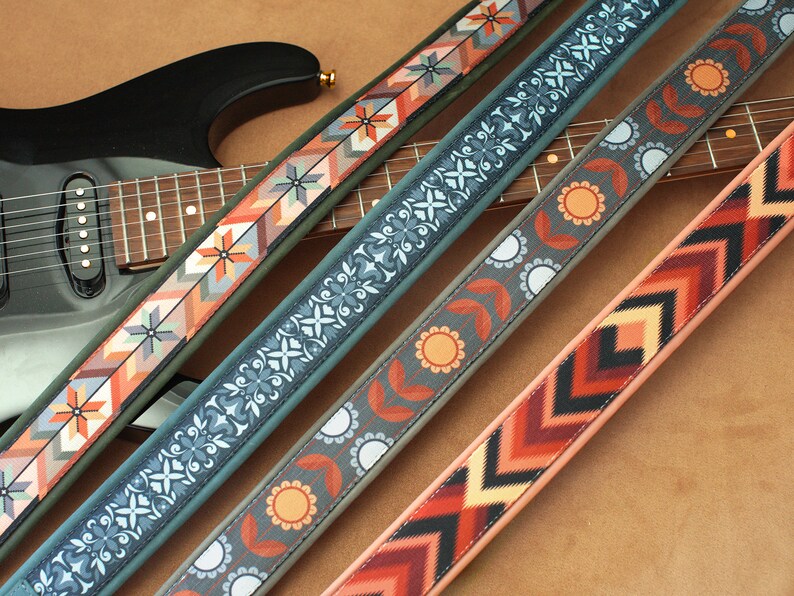 May include: Four guitar straps with different colorful patterns. The straps are made of fabric and have a woven design. The straps are laid out on a brown surface.