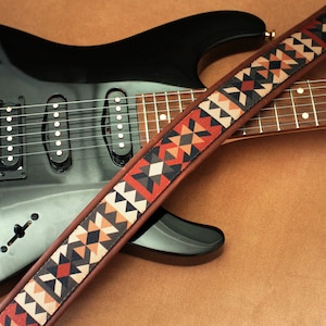 Puede incluir: Una guitarra el&eacute;ctrica negra con una correa de cuero marr&oacute;n con un patr&oacute;n geom&eacute;trico en tonos rojos, naranjas y negros.