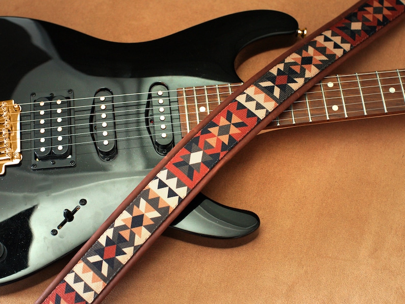 Puede incluir: Una guitarra el&eacute;ctrica negra con una correa de cuero marr&oacute;n con un patr&oacute;n geom&eacute;trico en tonos rojos, naranjas y negros.