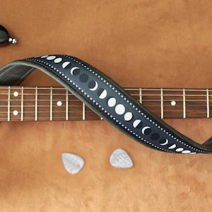 Puede incluir: Una correa de guitarra de cuero negro con un dise&ntilde;o de fases lunares blancas. La correa est&aacute; envuelta alrededor de una guitarra marr&oacute;n con un diapas&oacute;n de madera.