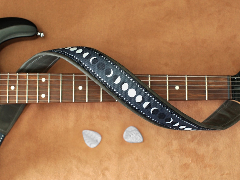 Puede incluir: Una correa de guitarra de cuero negro con un dise&ntilde;o de fases lunares blancas. La correa est&aacute; envuelta alrededor de una guitarra marr&oacute;n con un diapas&oacute;n de madera.