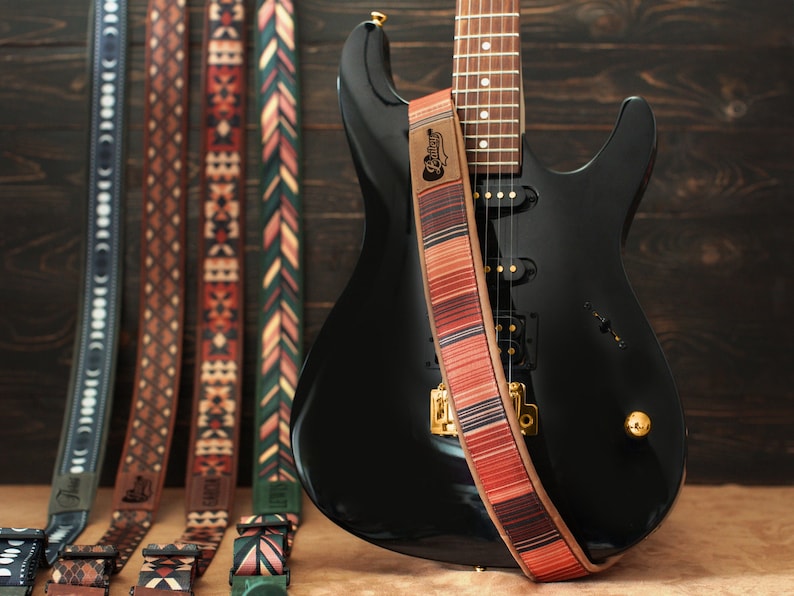 Puede incluir: Una guitarra el&eacute;ctrica negra con una correa de guitarra roja, blanca y marr&oacute;n con la palabra "Pelham" impresa en ella. La correa de guitarra est&aacute; colgando de la guitarra y est&aacute; enfocada. La guitarra est&aacute; en el fondo y est&aacute; desenfocada. Hay otras cuatro correas de guitarra en primer plano, cada una con un patr&oacute;n diferente.