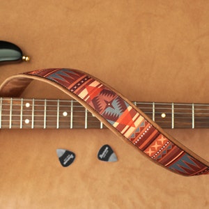 May include: A brown leather guitar strap with a colorful geometric pattern. The strap is wrapped around a black electric guitar with a wooden fretboard.