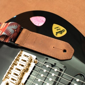 May include: A brown leather guitar strap with a patterned fabric strap attached. The strap is attached to a black electric guitar with gold hardware.