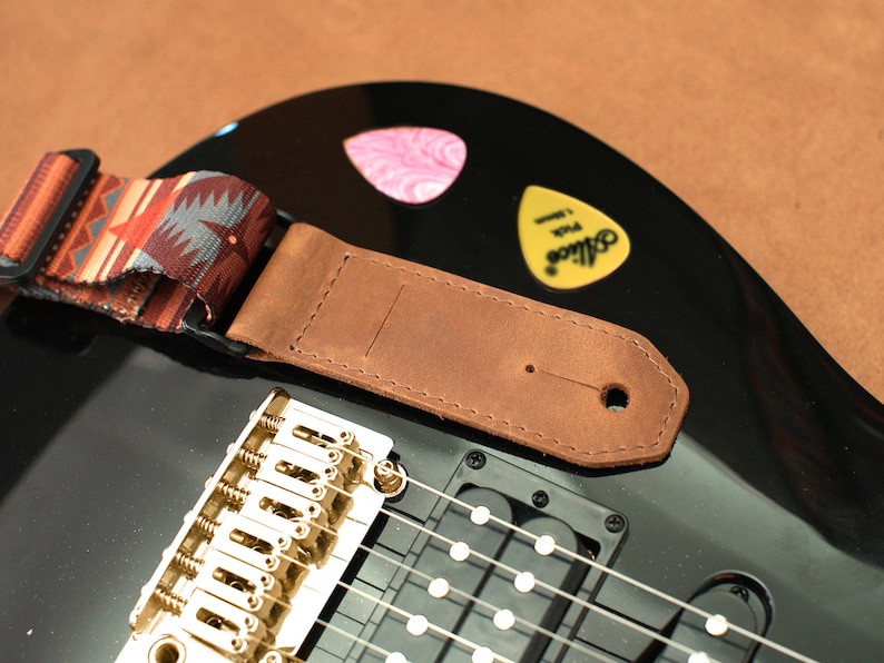 May include: A brown leather guitar strap with a patterned fabric strap attached. The strap is attached to a black electric guitar with gold hardware.