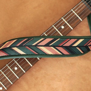 Puede incluir: Una correa de guitarra verde con un patr&oacute;n geom&eacute;trico rosa, beige y negro. La correa est&aacute; envuelta alrededor de un m&aacute;stil de guitarra marr&oacute;n.