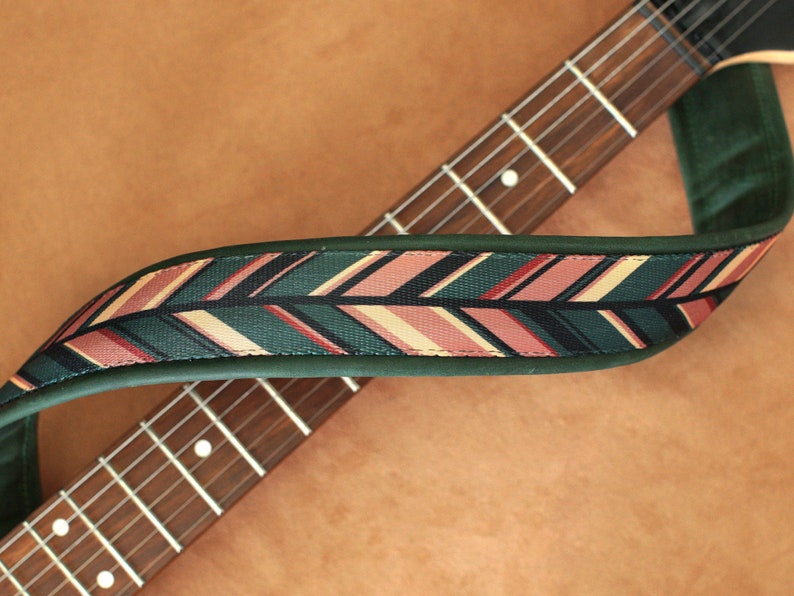 Puede incluir: Una correa de guitarra verde con un patr&oacute;n geom&eacute;trico rosa, beige y negro. La correa est&aacute; envuelta alrededor de un m&aacute;stil de guitarra marr&oacute;n.