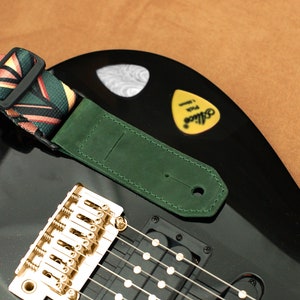 Puede incluir: Una guitarra el&eacute;ctrica negra con una correa de guitarra de cuero verde y una correa de guitarra con estampado colorido. La guitarra tiene herrajes dorados y una pastilla.