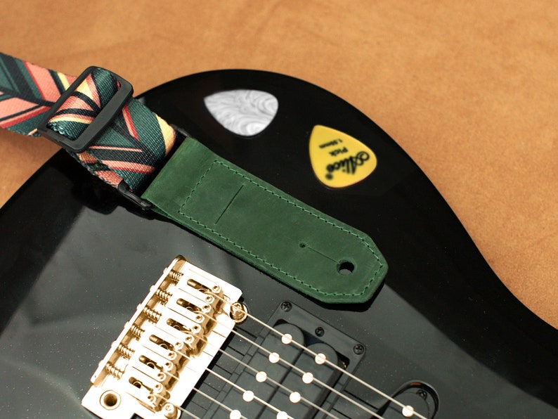 Puede incluir: Una guitarra el&eacute;ctrica negra con una correa de guitarra de cuero verde y una correa de guitarra con estampado colorido. La guitarra tiene herrajes dorados y una pastilla.