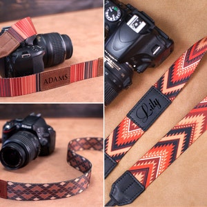 May include: Three camera straps with different patterns. The straps are made of fabric and have a leather patch with a name on it. The names are "Adams", "Lily", and "The Wanderer".
