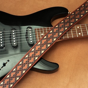 Puede incluir: Una guitarra el&eacute;ctrica negra con una correa de guitarra de patr&oacute;n marr&oacute;n y negro. La guitarra tiene tres pastillas y una clavija de afinaci&oacute;n dorada.