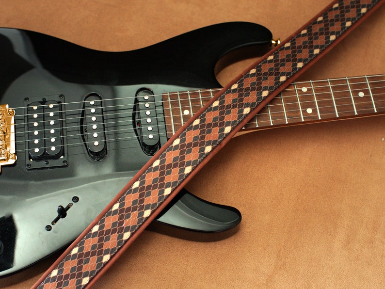 Puede incluir: Una guitarra el&eacute;ctrica negra con una correa de guitarra de patr&oacute;n marr&oacute;n y negro. La guitarra tiene tres pastillas y una clavija de afinaci&oacute;n dorada.
