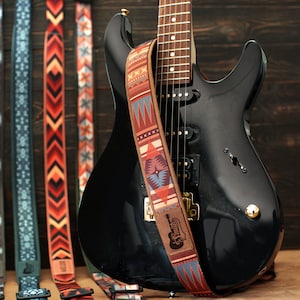 May include: Five guitar straps with different colorful patterns, including geometric designs and arrows. The straps are made of fabric and have leather ends. A black electric guitar with a brown wooden fretboard is in the background.