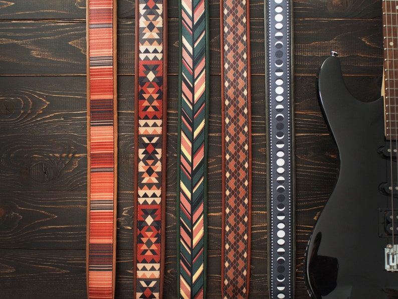 Puede incluir: Cinco correas de guitarra diferentes con varios patrones, incluyendo geom&eacute;tricos, chevron y fases lunares. Las correas son todas de cuero marr&oacute;n con dise&ntilde;os coloridos. Las correas est&aacute;n dispuestas sobre un fondo de madera oscura.