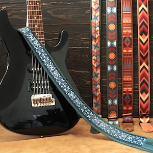 May include: A black electric guitar with a green and white patterned guitar strap. The strap has the word "Amelia" printed on it. Four other guitar straps with different patterns are visible in the background.