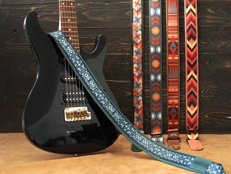 May include: A black electric guitar with a green and white patterned guitar strap. The strap has the word "Amelia" printed on it. Four other guitar straps with different patterns are visible in the background.