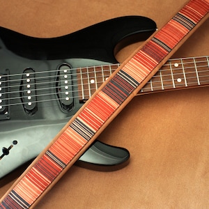 Puede incluir: Una guitarra el&eacute;ctrica negra con una correa de guitarra a rayas marrones y rojas. La guitarra tiene tres botones plateados y un interruptor de palanca plateado.