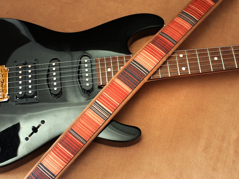Puede incluir: Una guitarra el&eacute;ctrica negra con una correa de guitarra a rayas marrones y rojas. La guitarra tiene tres botones plateados y un interruptor de palanca plateado.