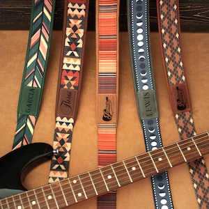 Puede incluir: Seis correas de guitarra con diferentes patrones y nombres personalizados, incluyendo "Garcia", "Flores", "James", "Lewis" y "Bailey". Las correas est&aacute;n dispuestas sobre una superficie marr&oacute;n con una guitarra encima.