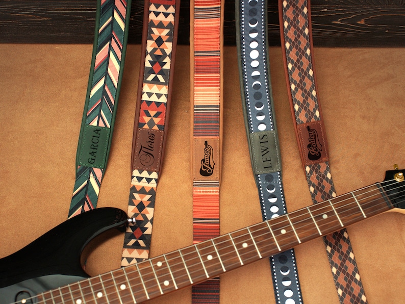 Puede incluir: Seis correas de guitarra con diferentes patrones y nombres personalizados, incluyendo "Garcia", "Flores", "James", "Lewis" y "Bailey". Las correas est&aacute;n dispuestas sobre una superficie marr&oacute;n con una guitarra encima.