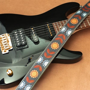 May include: A black electric guitar with a gold bridge and a patterned guitar strap with orange, blue, and white accents.