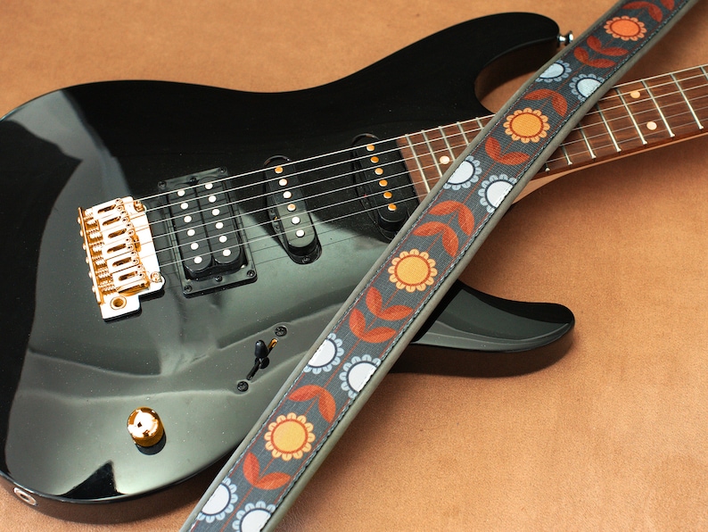 May include: A black electric guitar with a gold bridge and a patterned guitar strap with orange, blue, and white accents.