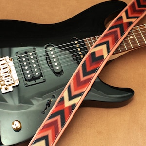 May include: A black electric guitar with a gold bridge and a patterned guitar strap with red, black, and cream chevron designs.