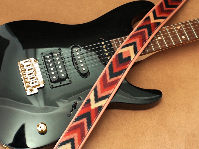 May include: A black electric guitar with a gold bridge and a patterned guitar strap with red, black, and cream chevron designs.